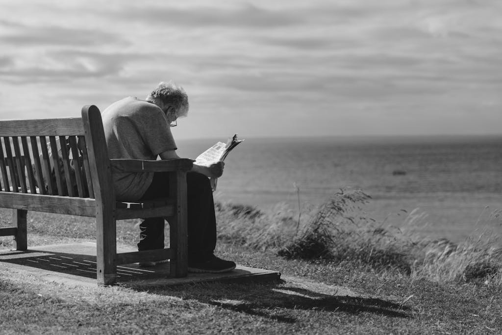A contemplative scene of an elderly man reading a newspaper on a seaside bench with cloudy skies.
