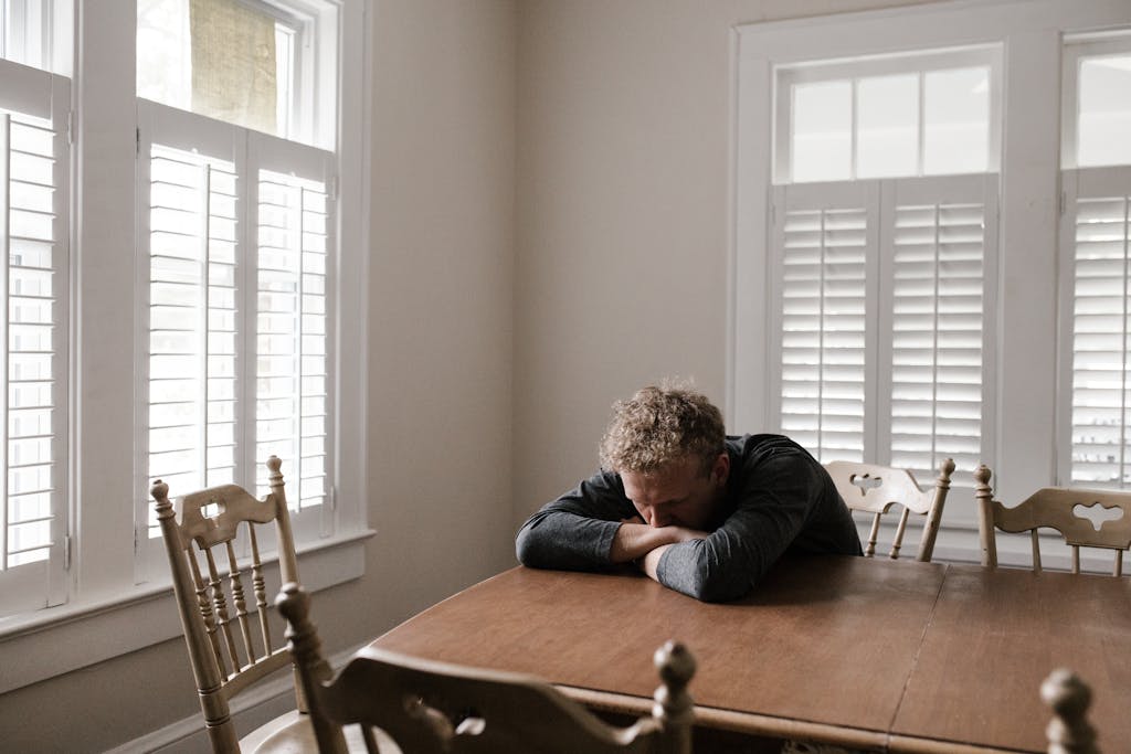 An adult man with emotions of sadness and anxiety sitting alone at a wooden table near windows.